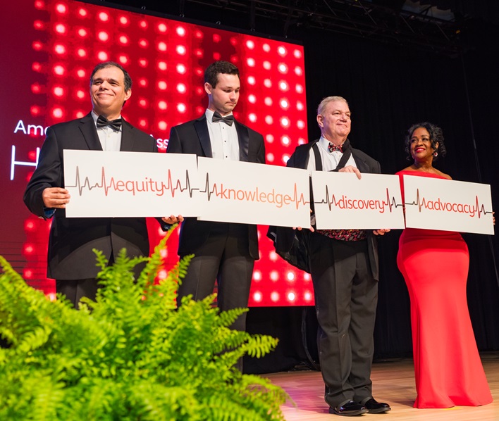 Photo of four people holding up a banner reading equity, knowledge, discovery, and advocacy with each word separated by an EKG reading